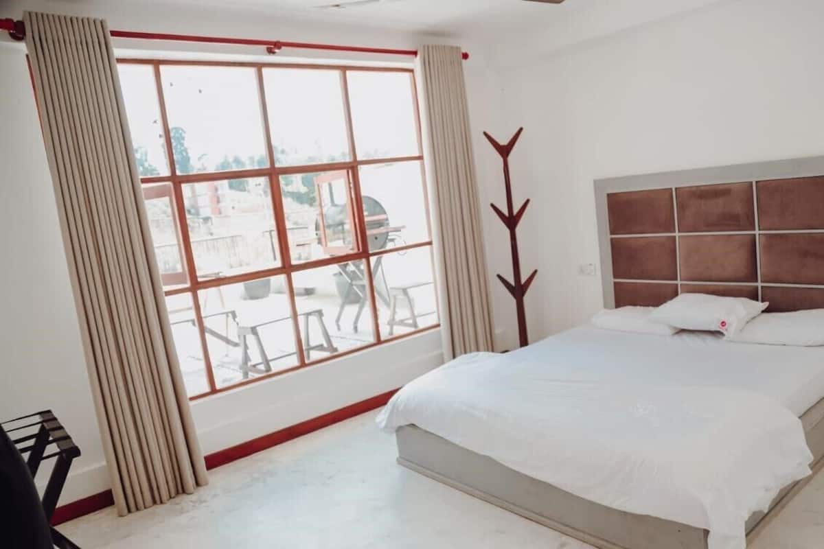 Une chambre avec une grande fenêtre, un lit recouvert de draps blancs et une tête de lit à motifs géométriques.