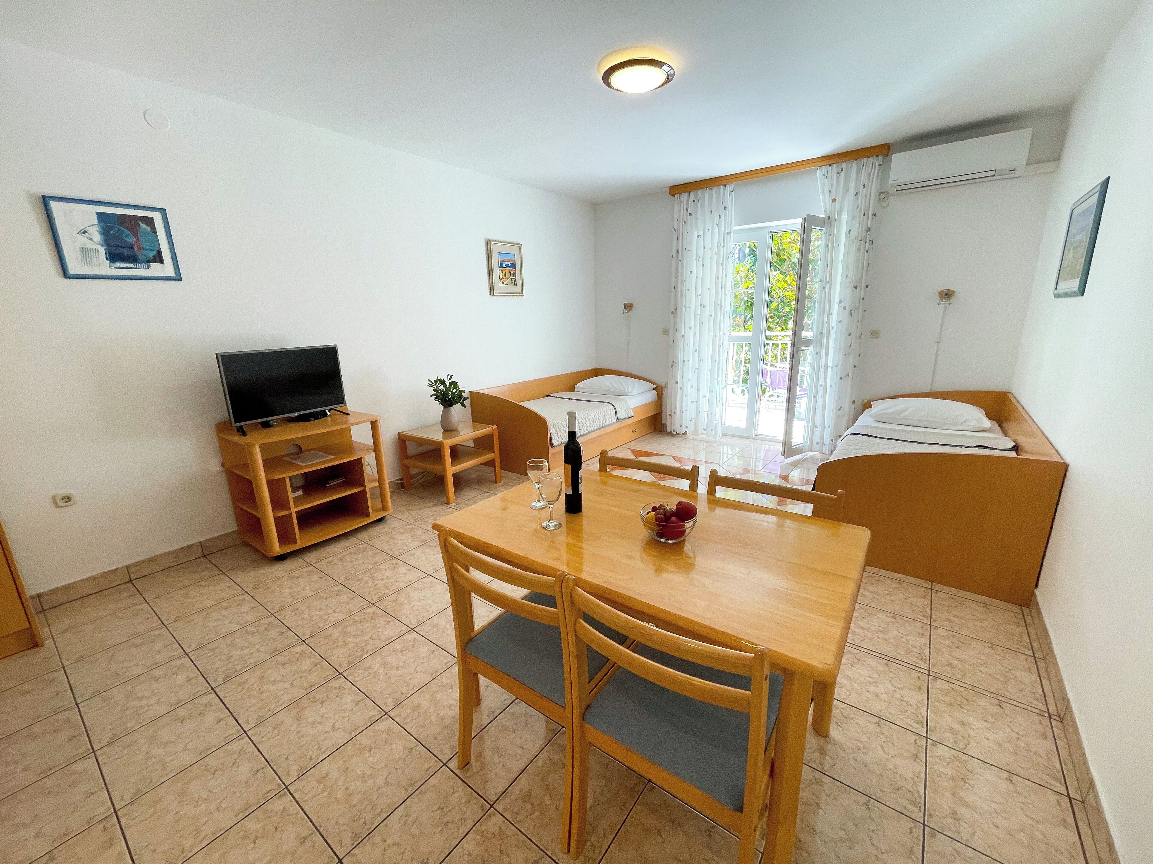 A small, tidy room with a wooden table, two chairs, a TV on a stand, a bed with a white mattress, and a window with curtains.