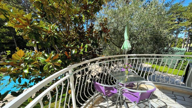 A balcony with a glass table and purple chairs, surrounded by greenery and a pool. - Apartman Villa Ljiljana 2 (Jadranovo)