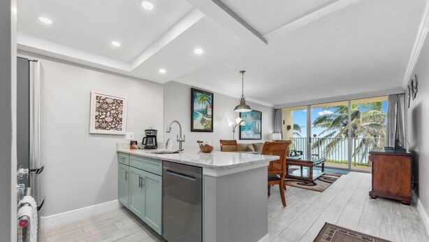 A modern kitchen with a central island, a dining area with wooden chairs, and a view of palm trees through large windows.