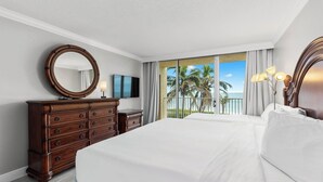 A hotel room with a large bed, a wooden dresser, a round mirror, a flat-screen TV, and a balcony with a view of palm trees and the ocean.