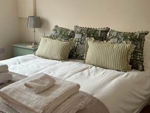 Una cama bien tendida con sábanas blancas y almohadas de estampado verde, una lámpara en una mesita de noche y toallas dobladas en una silla de mimbre.