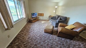 Ein Wohnzimmer mit grauem Sofa, braunem Ledersitzhocker und einem Fernseher auf einem Holzgestell.