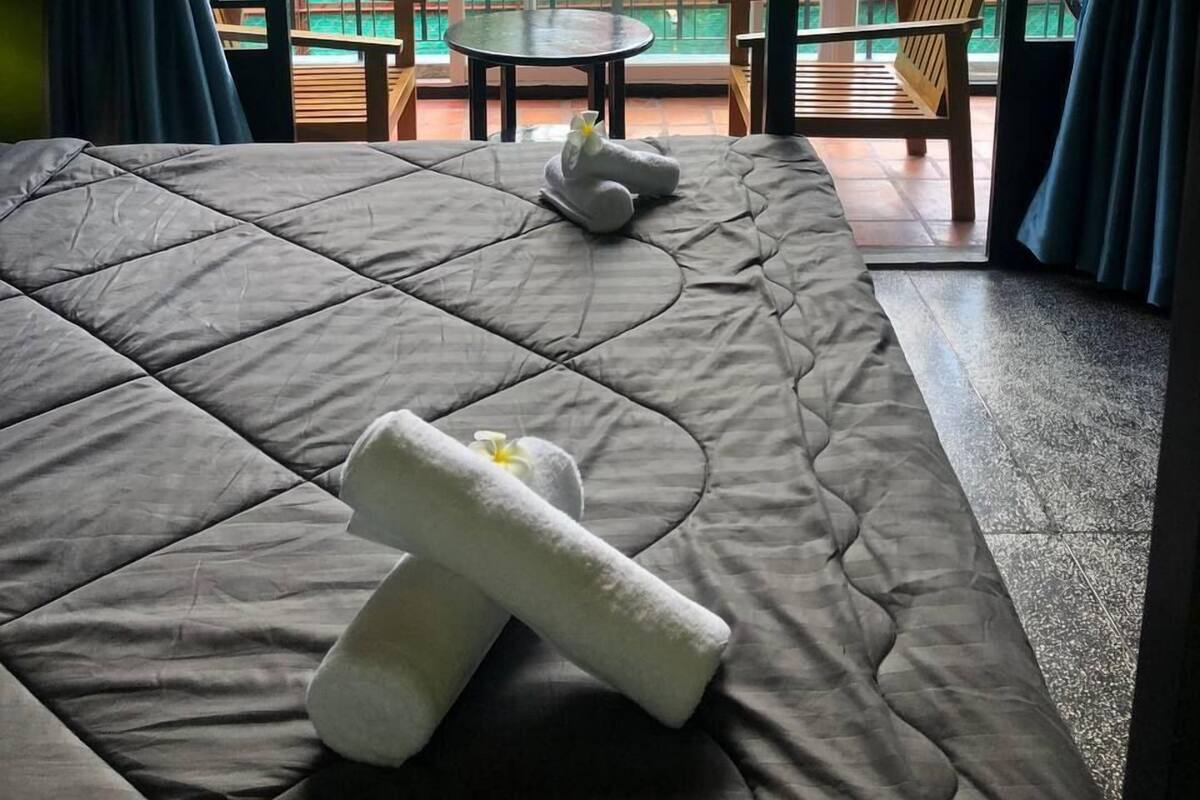 A room with a bed, a small table, and a chair. There is a balcony with a view of a house and greenery.