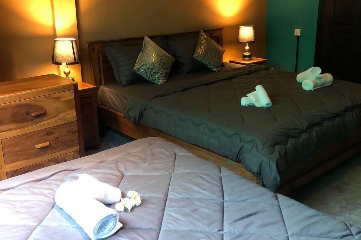 A hotel room with two beds, each with a quilted mattress and pillows. There are two bedside tables with lamps, a wooden dresser, and a green wall.