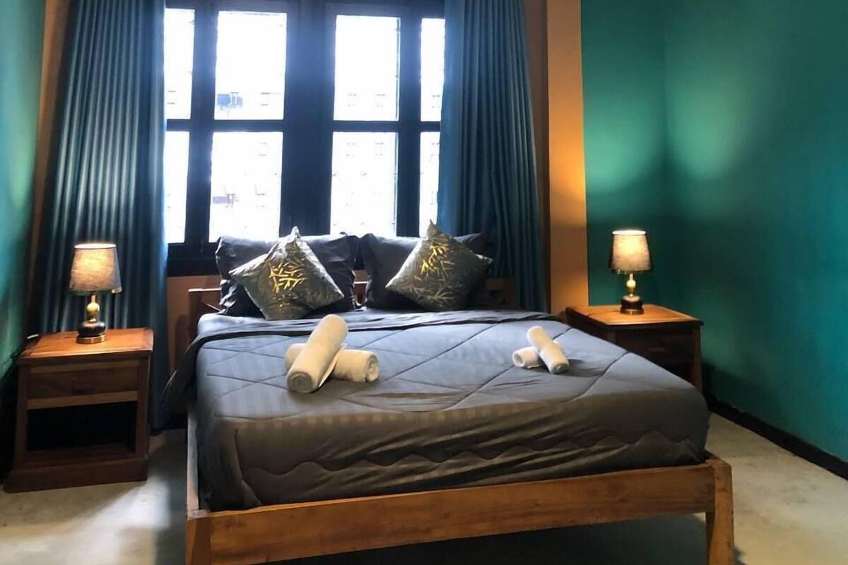 A bedroom with a bed, two bedside tables, a hanging light fixture, an air conditioning unit, and a window with curtains.