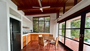 Una cocina con mesa y sillas de madera, un refrigerador negro y ventanas amplias con vistas a la vegetación.