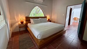 Un dormitorio con una cama grande, dos mesitas de noche con lámparas, una ventana con vista a follaje y un ventilador de techo.