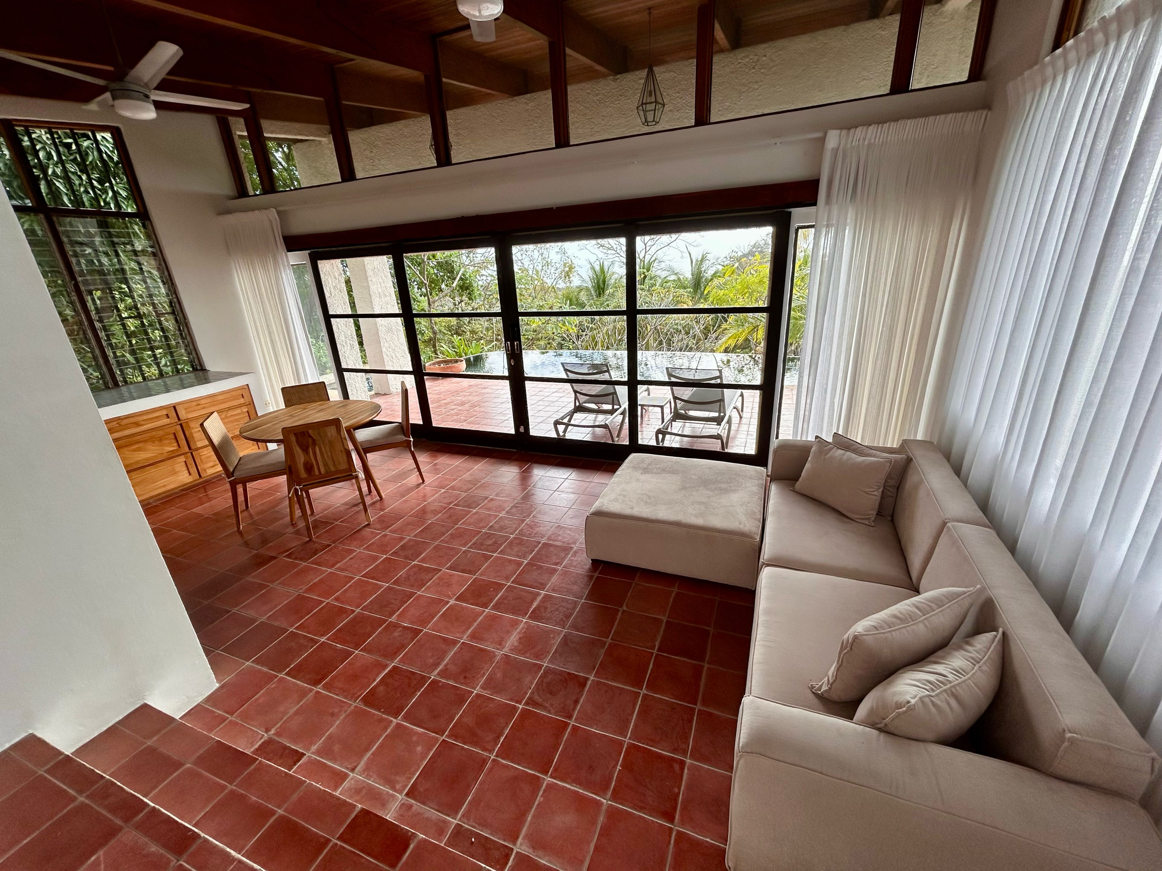 A spacious living area with a sofa, a dining table with chairs, and a view through large windows to an outdoor patio.