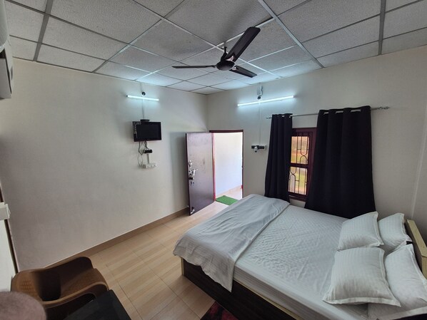 A hotel room with a bed, a ceiling fan, a flat-screen TV mounted on the wall, and a window with curtains.