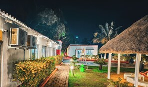 A nighttime view of a resort with a thatched-roof structure, a paved pathway, and a building with a red door.