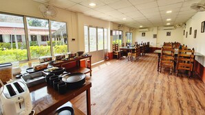Un comedor con mesas y sillas de madera, un bufé con distintos alimentos y amplias ventanas que ofrecen vistas al exterior con vegetación.