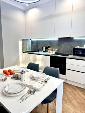 A modern kitchen with white cabinetry, a built-in oven, and a dining area set with plates, cutlery, and a fruit bowl.