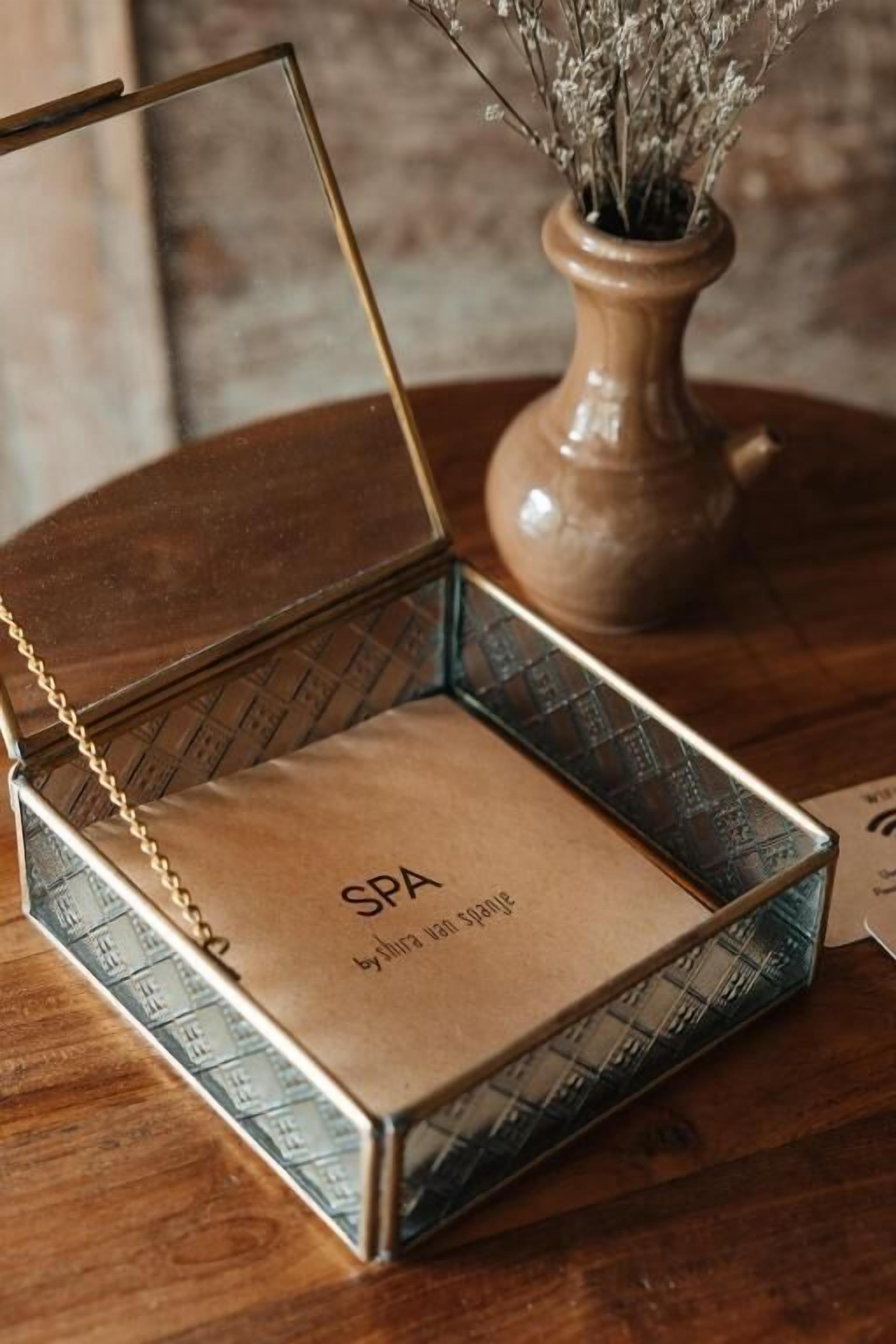 A spa gift box with a gold chain and a brown interior, placed on a wooden table next to a vase with dried flowers.