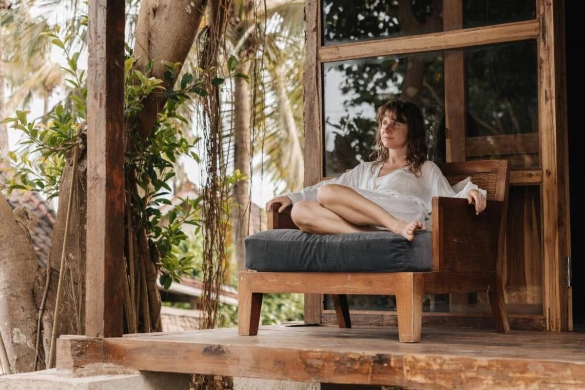 A person sitting on a wooden bench with a cushion, outdoors near a wooden structure with a window.