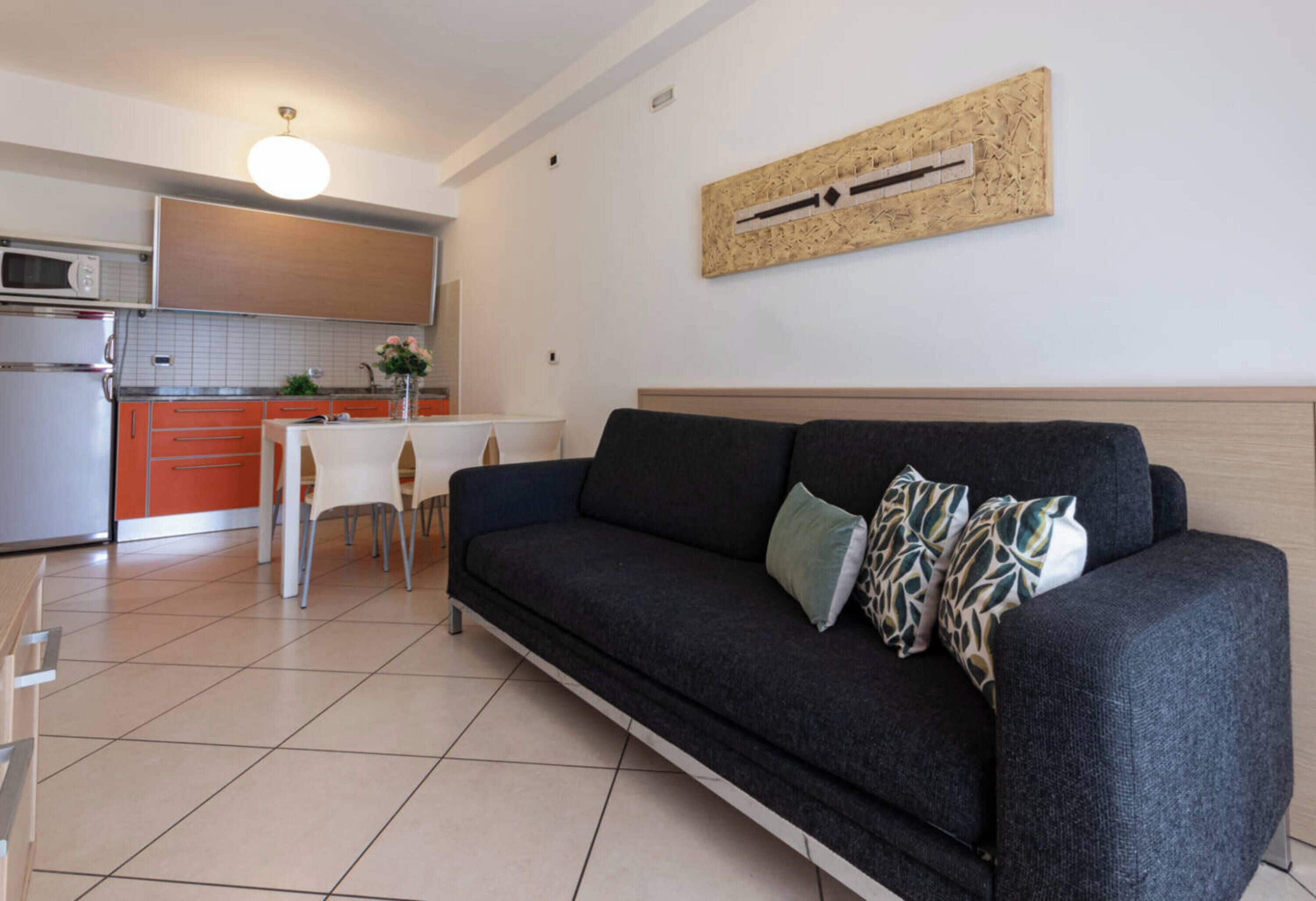 A modern living room with a dark grey sofa, a small dining table, and a kitchen area in the background.