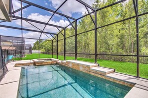 A swimming pool with a glass enclosure, surrounded by a wooden deck and a view of a green lawn and trees. - 4736 Family Home Near Disney Sea World (Kissimmee)