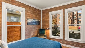 Una habitación con pared de ladrillo, cama con colcha azul, ventana con vista a un edificio y un cuadro en la pared.