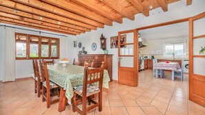 A spacious dining area with wooden chairs and a table, a kitchen with modern appliances, and a view through an open door. - Villa Can Paxaco - By Dream Villas Pollensa (Alcudia)