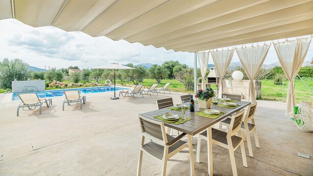 An outdoor patio with a pool, dining table, and chairs, surrounded by a covered area and a view of the mountains. - Villa Can Paxaco - By Dream Villas Pollensa (Alcudia)