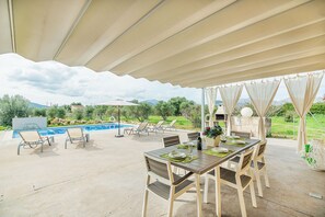 An outdoor patio with a pool, dining table, and chairs, surrounded by a covered area and a view of the mountains.