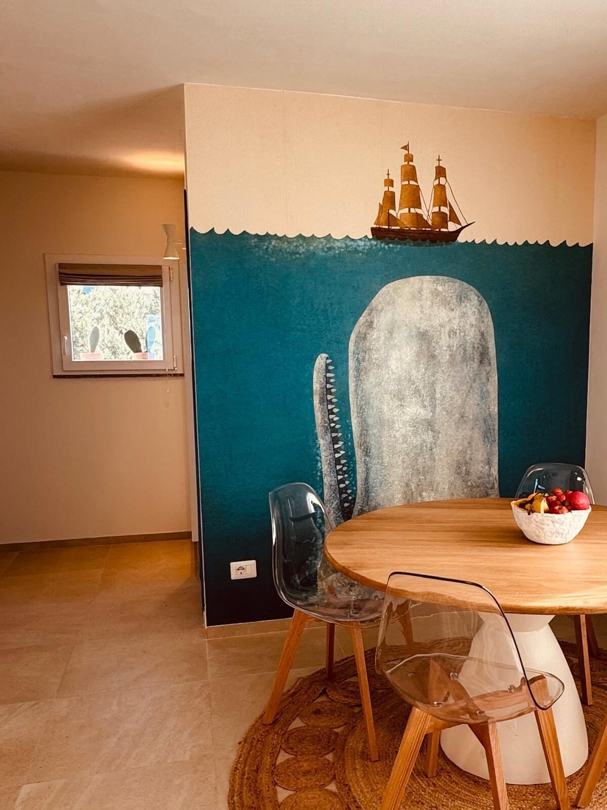 Ein Essbereich mit einem runden Holztisch, transparenten Glasstühlen, einem gewebten Teppich und einem Wandbild mit einem Schiff und einem Wal.