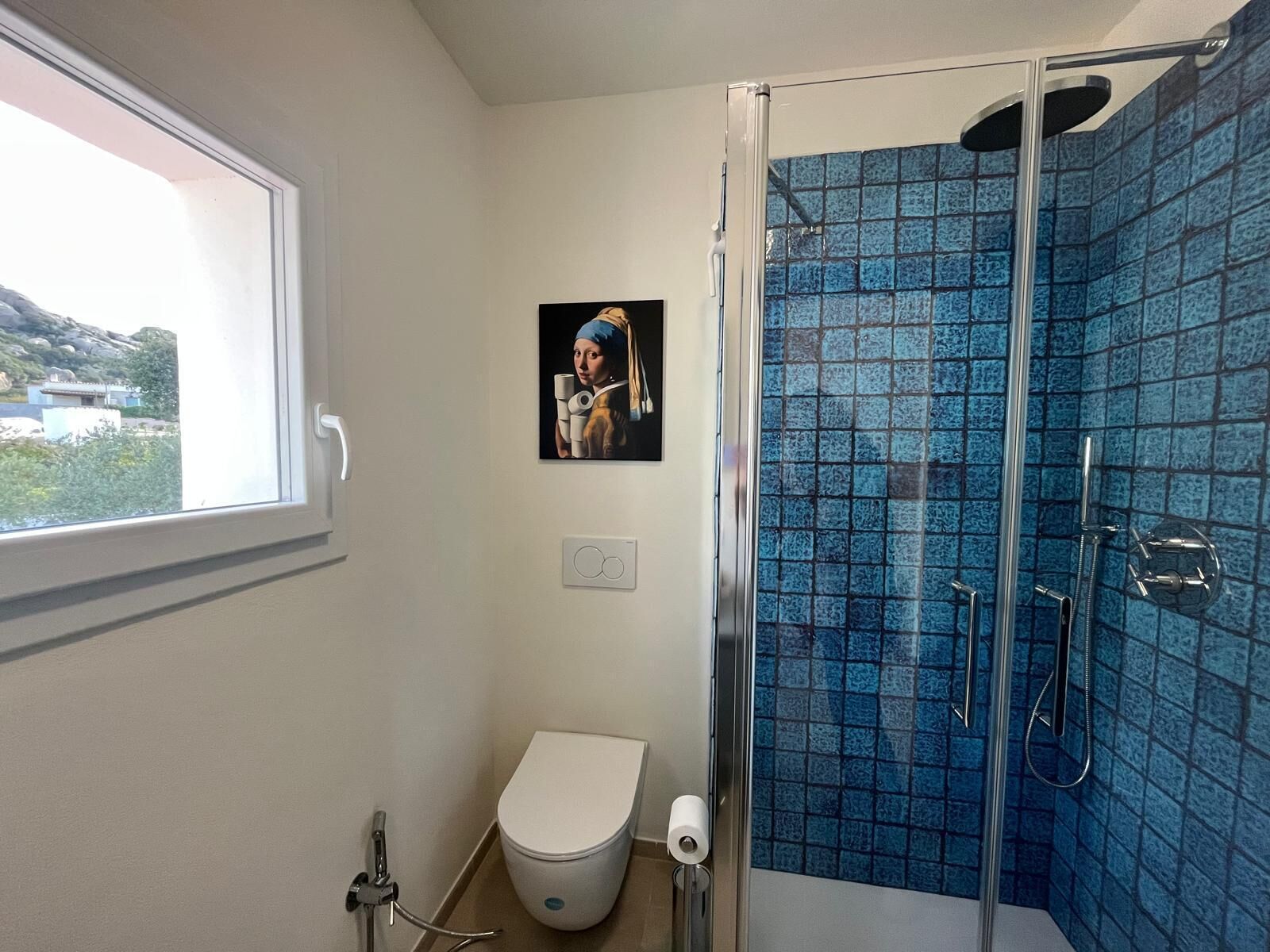 A bathroom with a toilet, a shower with blue tiles, a window, and a painting on the wall.
