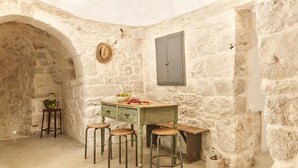 A rustic stone-walled room with a wooden bar and stools, a small table with fruit, and a hat on the wall. - Trullo dei Noci con Piscina Privata (Ceglie Messapica)