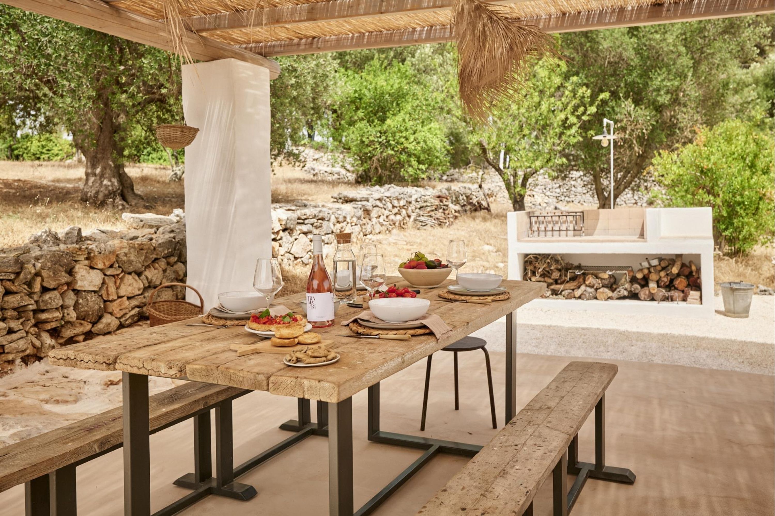 Un área de comedor al aire libre con una mesa de madera rústica dispuesta para una comida, rodeada de muros de piedra y árboles.