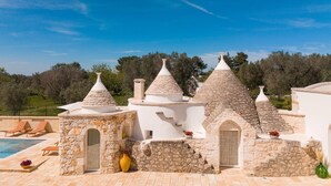 A villa with stone and white structures, a swimming pool, and a view of the countryside.