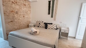 A bedroom with a stone wall, a bed with a beige bedspread, patterned pillows, and a white nightstand with a lamp. - Villa Vittoria in Ostuni (Ostuni)