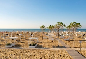 A beach with white lounge chairs and tables, a wooden walkway, and trees. - Charming Apartment in the Center of Bibione (San Michele al Tagliamento)