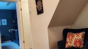 A room with a dark blue armchair, a dartboard, and a kitchen area visible through an open door. - Chic & Modern Escape (Salford)