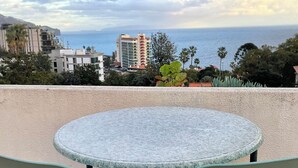 Mesa redonda con dos sillas en un balcón con vista a un paisaje urbano costero, con edificios y palmeras.