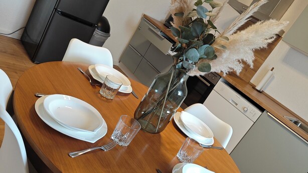 A dining table set with plates, glasses, and cutlery, surrounded by white chairs. A black refrigerator and a white dishwasher are visible in the background. - Your vacation home (Andernach)