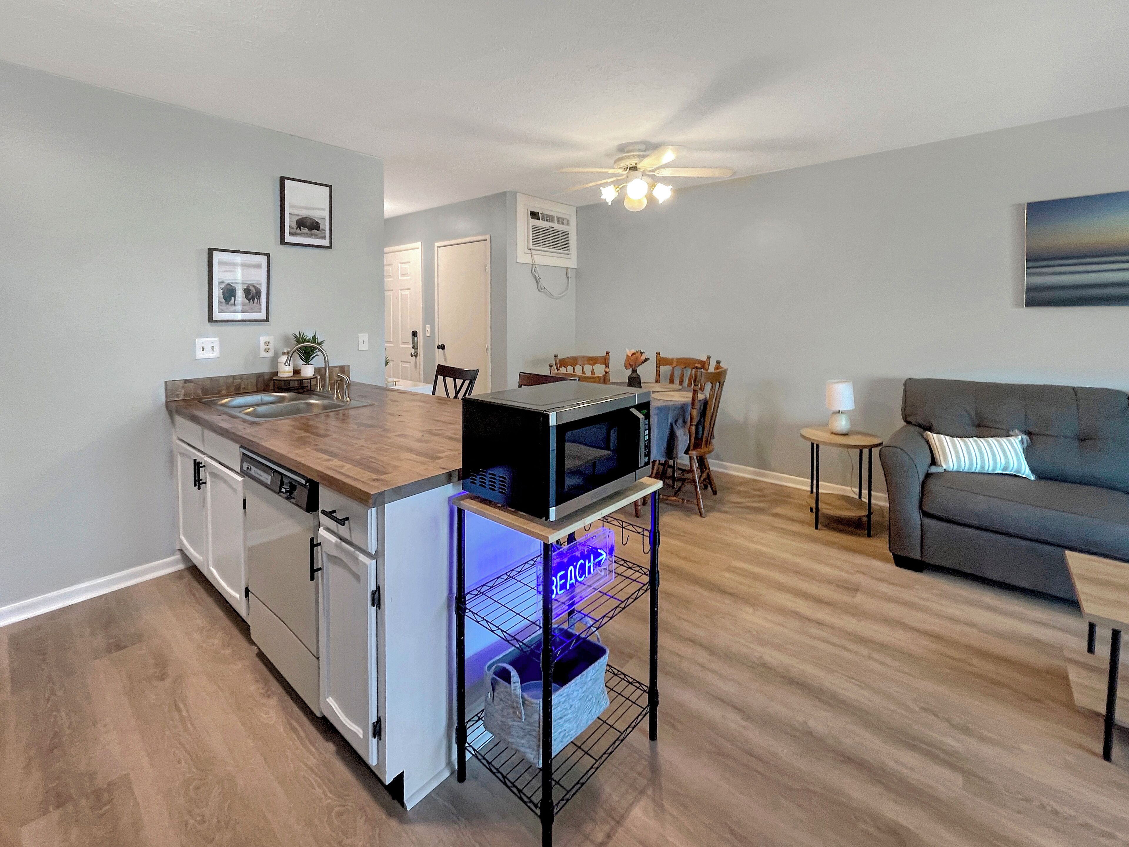 Una cocina moderna con lavavajillas integrado, microondas y un refrigerador pequeño. Hay un comedor con mesa y sillas, y una sala de estar con sofá y lámpara.