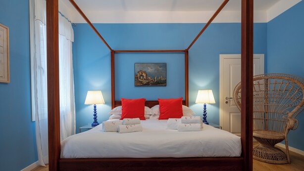 A four-poster bed with white linens and red pillows, a painting on the wall, and a wicker chair.