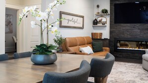 Un salon moderne avec une cheminée, un coin repas avec une grande table et des chaises, et un canapé agrémenté d’un coussin vert.