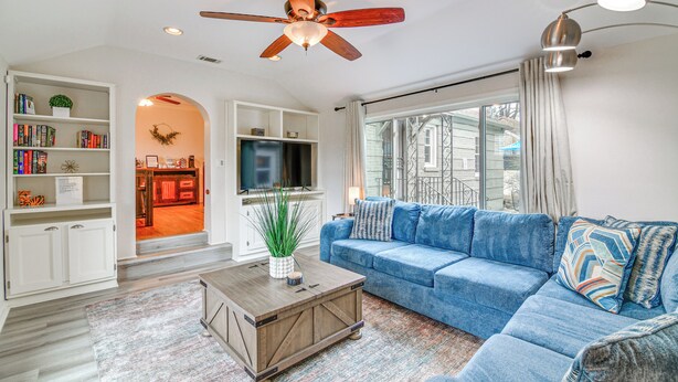 A living room with a blue sofa, a wooden coffee table, and a ceiling fan. There is a bookshelf with books and a TV mounted on the wall. - Spacious Memphis Retreat: 5 Mi to Downtown Beats! (Memphis)