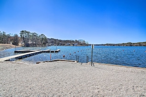 A pebbled beach area with a dock extending into a calm lake, surrounded by trees and buildings in the distance.