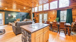 L’intérieur d’une cabane en bois est agrémenté d’un mur d’accent en pierre, d’un îlot central avec une cuisinière, d’un coin repas avec des chaises en bois et d’une grande fenêtre avec des rideaux.