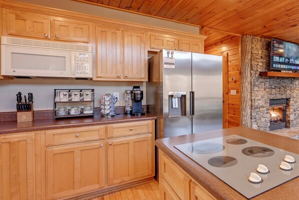 A kitchen with wooden cabinets, a stainless steel refrigerator, a microwave, a coffee maker, and a stone fireplace.
