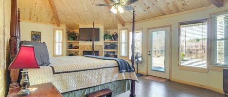 A bedroom with a wooden ceiling, a bed with a quilt, a nightstand, a red lamp, a wooden bench, a ceiling fan, and a window with blinds.