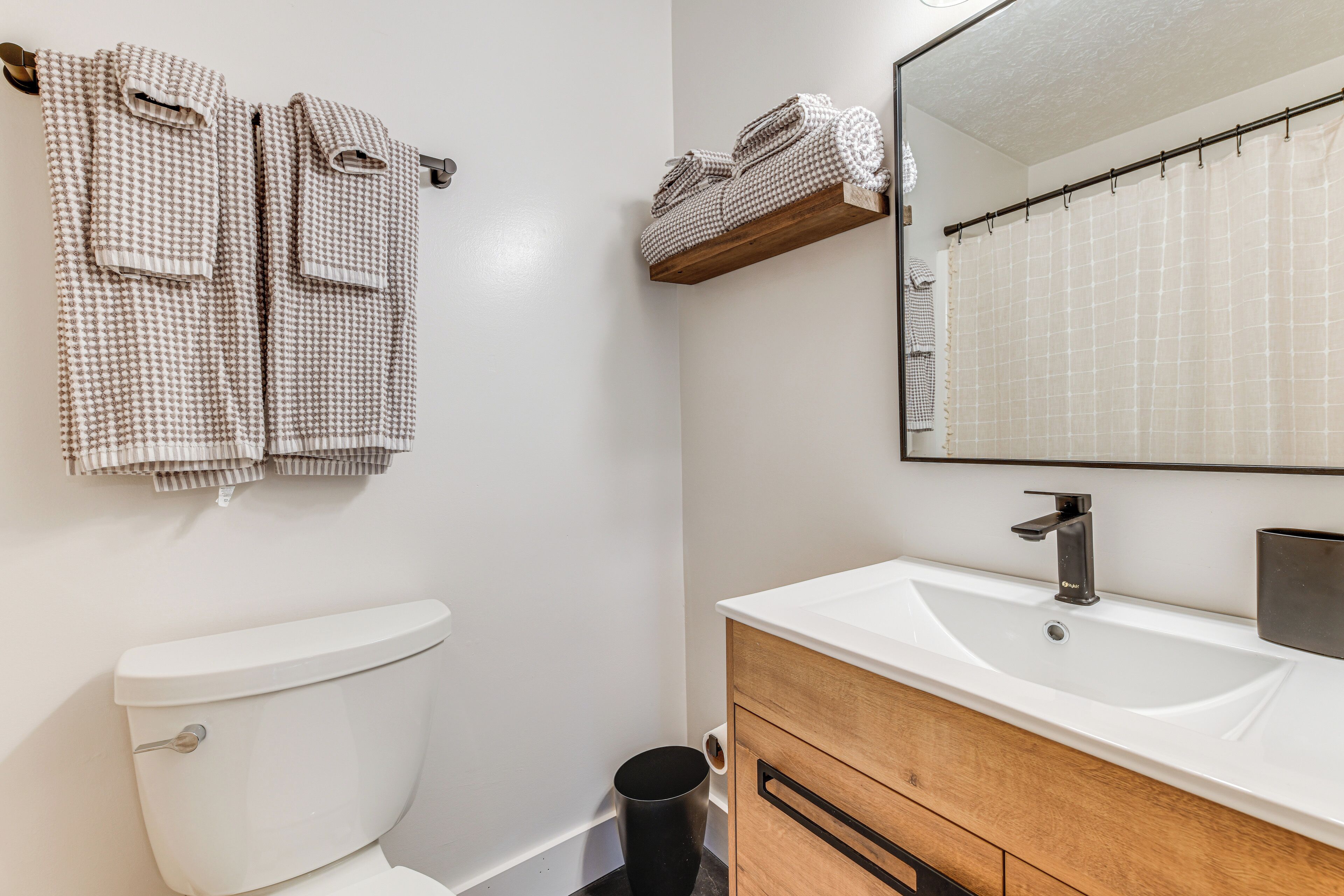 Une salle de bain avec des toilettes blanches, un meuble-lavabo en bois avec un évier et un miroir au-dessus du meuble. Des serviettes sont suspendues à un porte-serviettes et il y a une étagère avec des serviettes pliées.