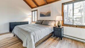 Quarto com cama grande, vigas de madeira no teto, radiador e janela com vista para árvores.