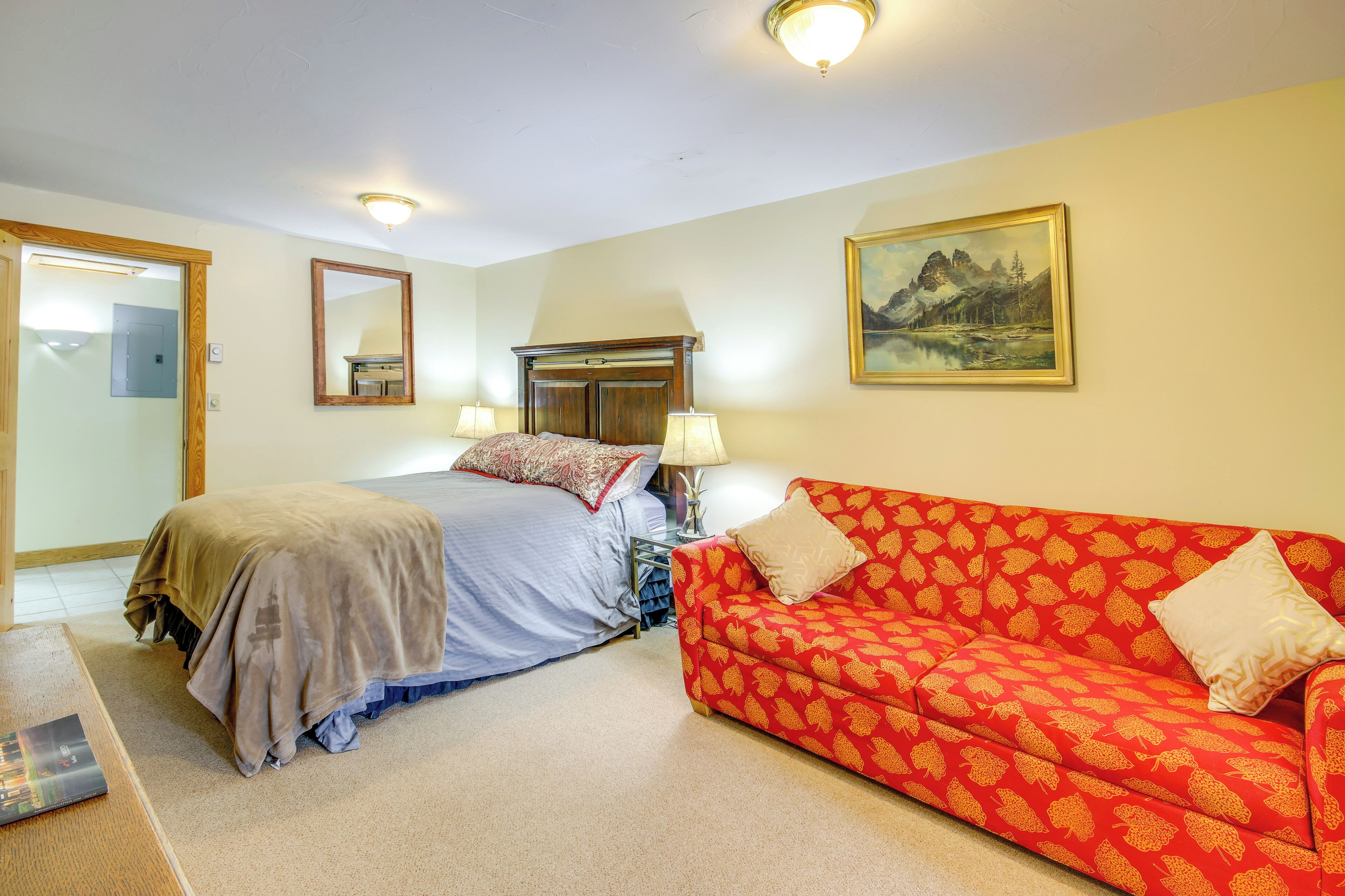 A bedroom with a bed, a red patterned sofa, a painting on the wall, and a mirror.