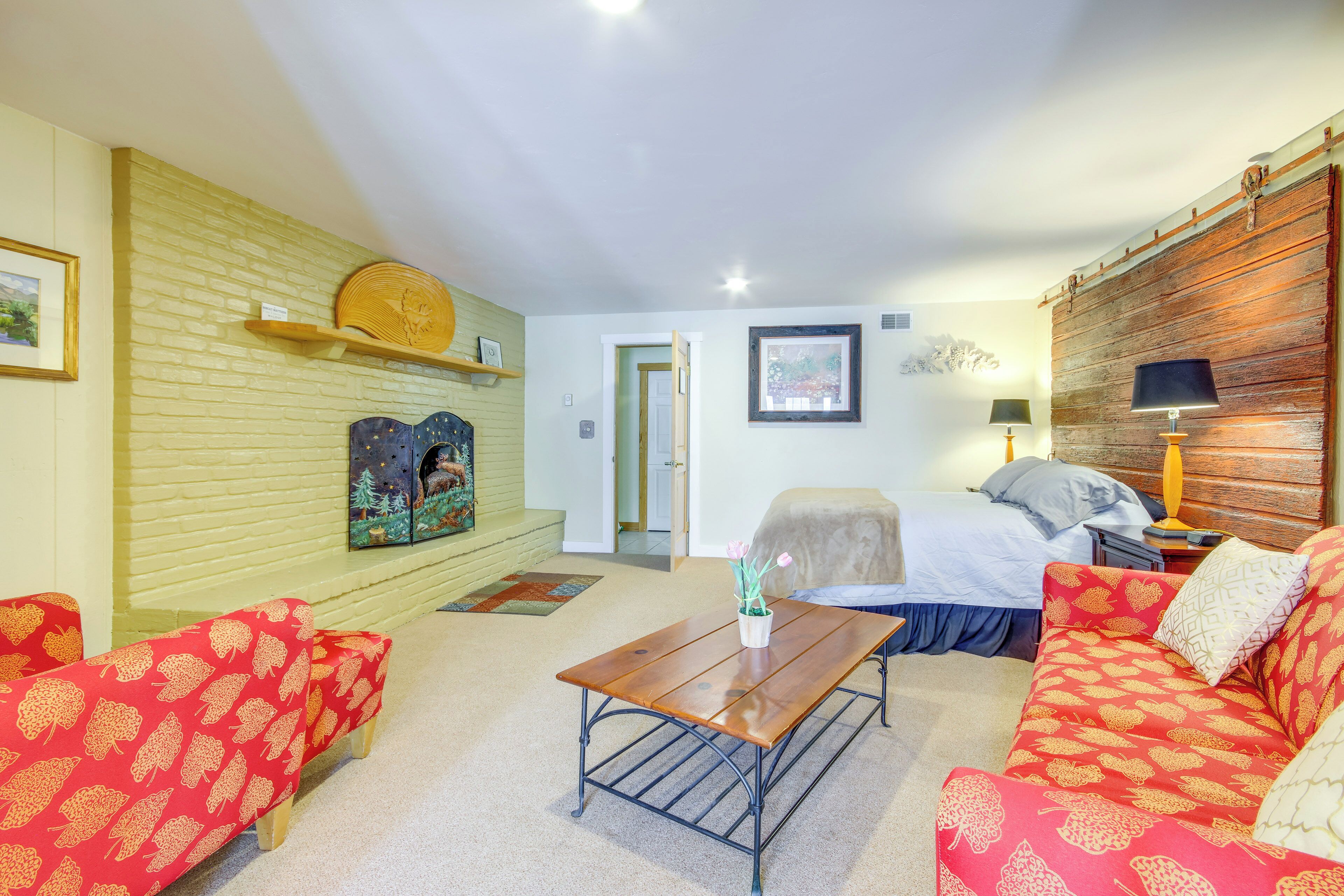 A room with a bed, two red patterned sofas, a wooden coffee table, and a wall-mounted shelf with a decorative item.