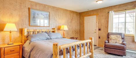 Une chambre avec un lit en bois, deux lampes de chevet, un ventilateur de plafond, une grande fenêtre et un fauteuil avec un coussin.