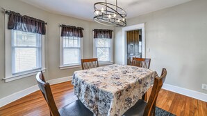A dining area with a table covered in a floral tablecloth, surrounded by wooden chairs, and three windows with brown curtains. - 1 Mi to Dtwn Binghamton: Apt w/ Covered Balcony! (Binghamton)