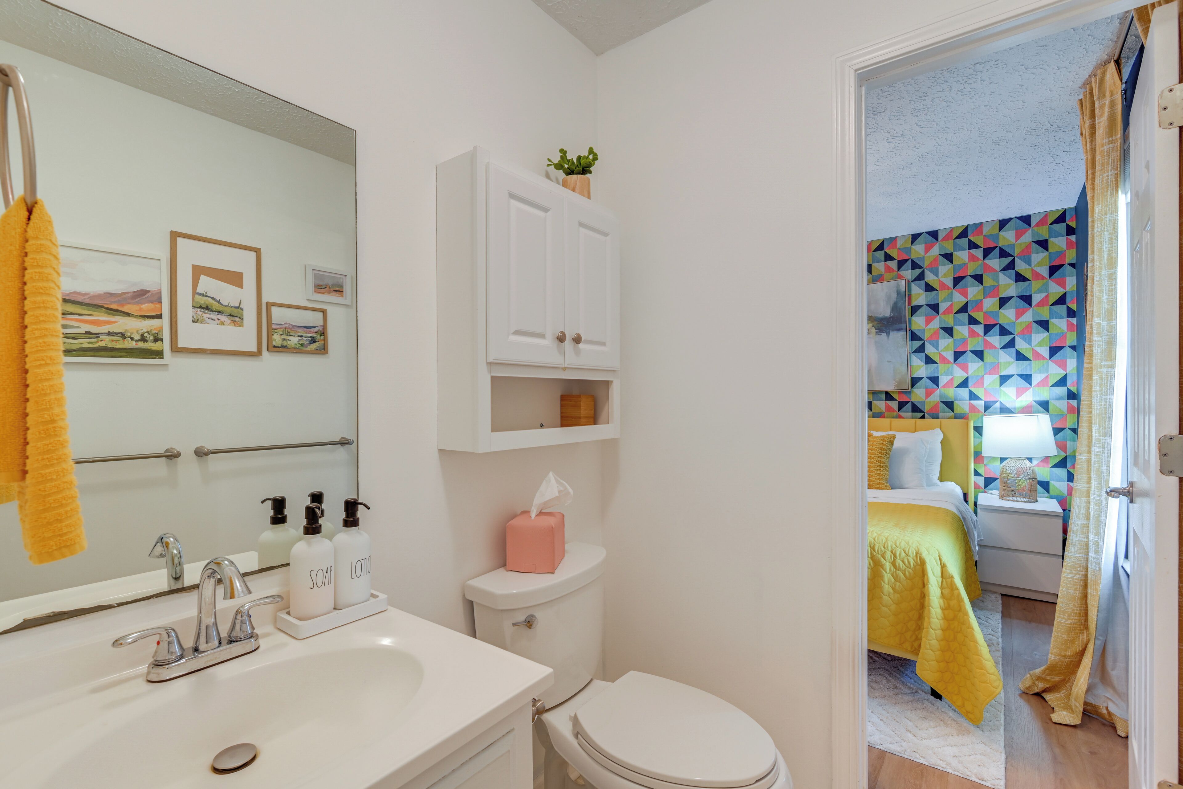 A bathroom with a sink, mirror, and toilet. There is a bed with a yellow bedspread in the adjacent room.